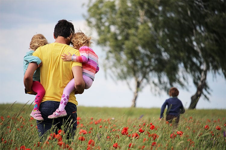 father carrying two daughters as he walks after his son through field of flowers