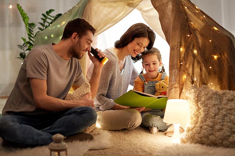mother, father, and daughter inside a blanket fort reading a book together