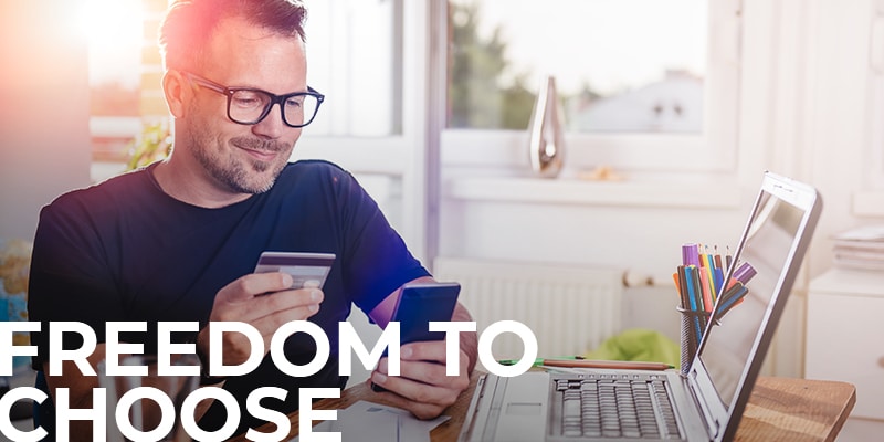 man sitting at his home desk, credit card in one hand, smartphone in the other, with a text overlay: Freedom To Choose