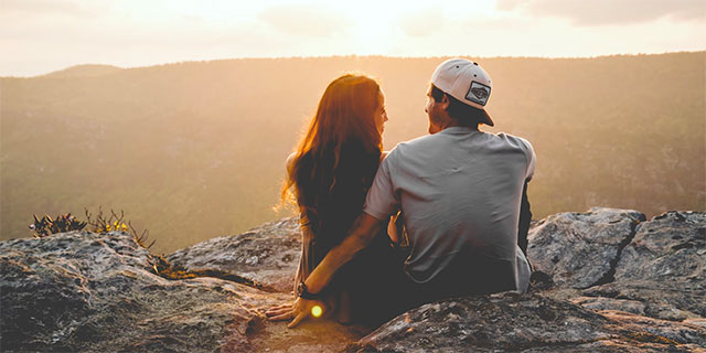 A husband and wife watching the sun set from a mountain top.