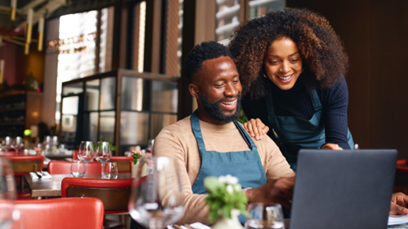 A Black woman and man inside their business, a restaurant, looking at their computer.